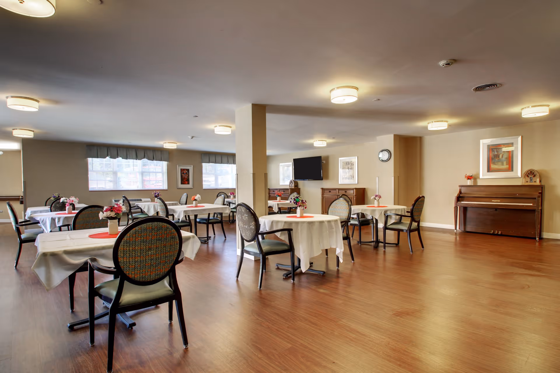A spacious dining room with several round tables covered with white tablecloths, each adorned with a small vase of flowers. The room has wooden flooring, beige walls, multiple ceiling lights, a wall-mounted TV, a clock, framed artwork, and a piano against one wall.