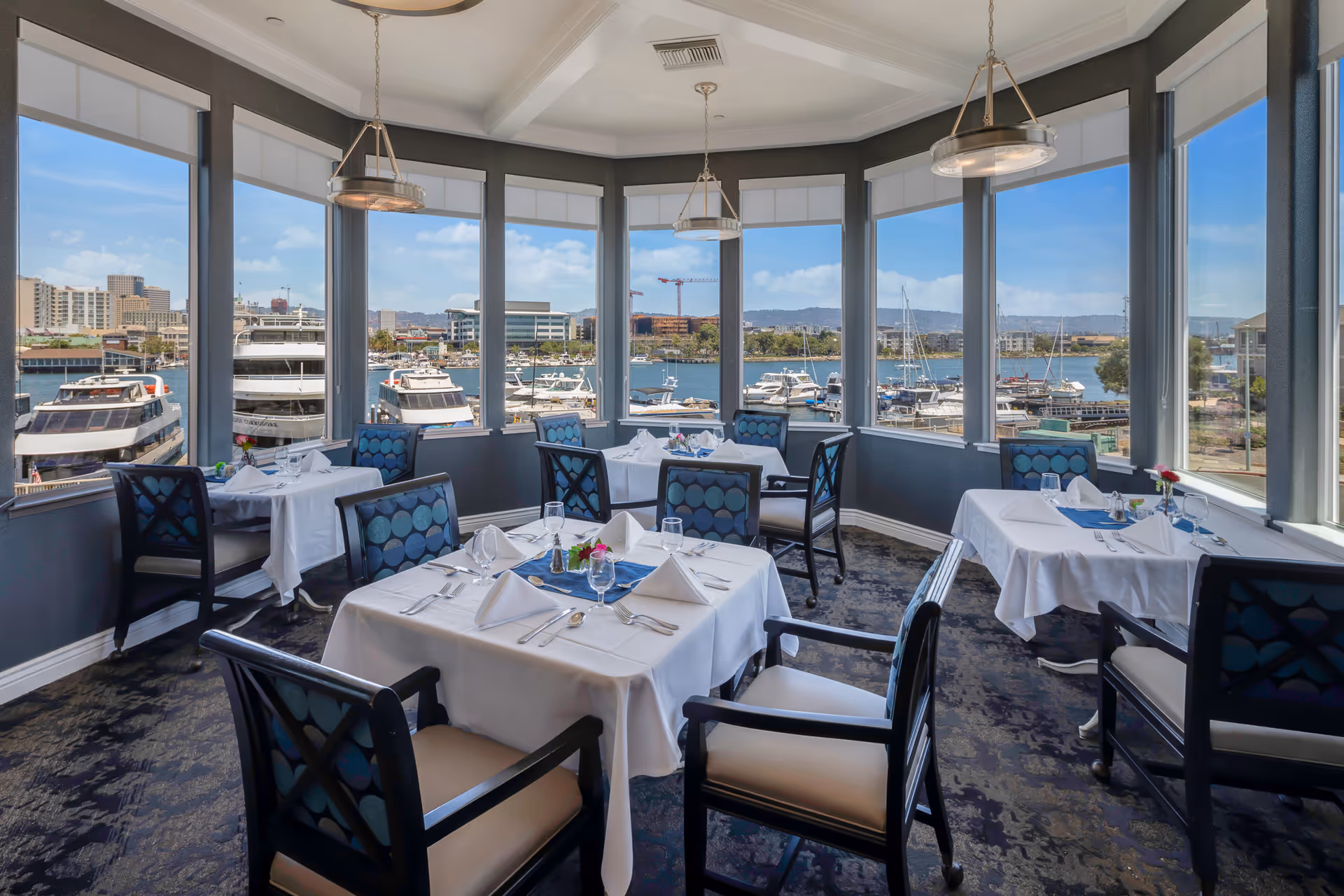Dining room with several tables covered in white tablecloths, set with napkins, glasses, and silverware. The room has large windows offering a panoramic view of a marina with boats and buildings in the background. The chairs have dark frames with blue patterned cushions.