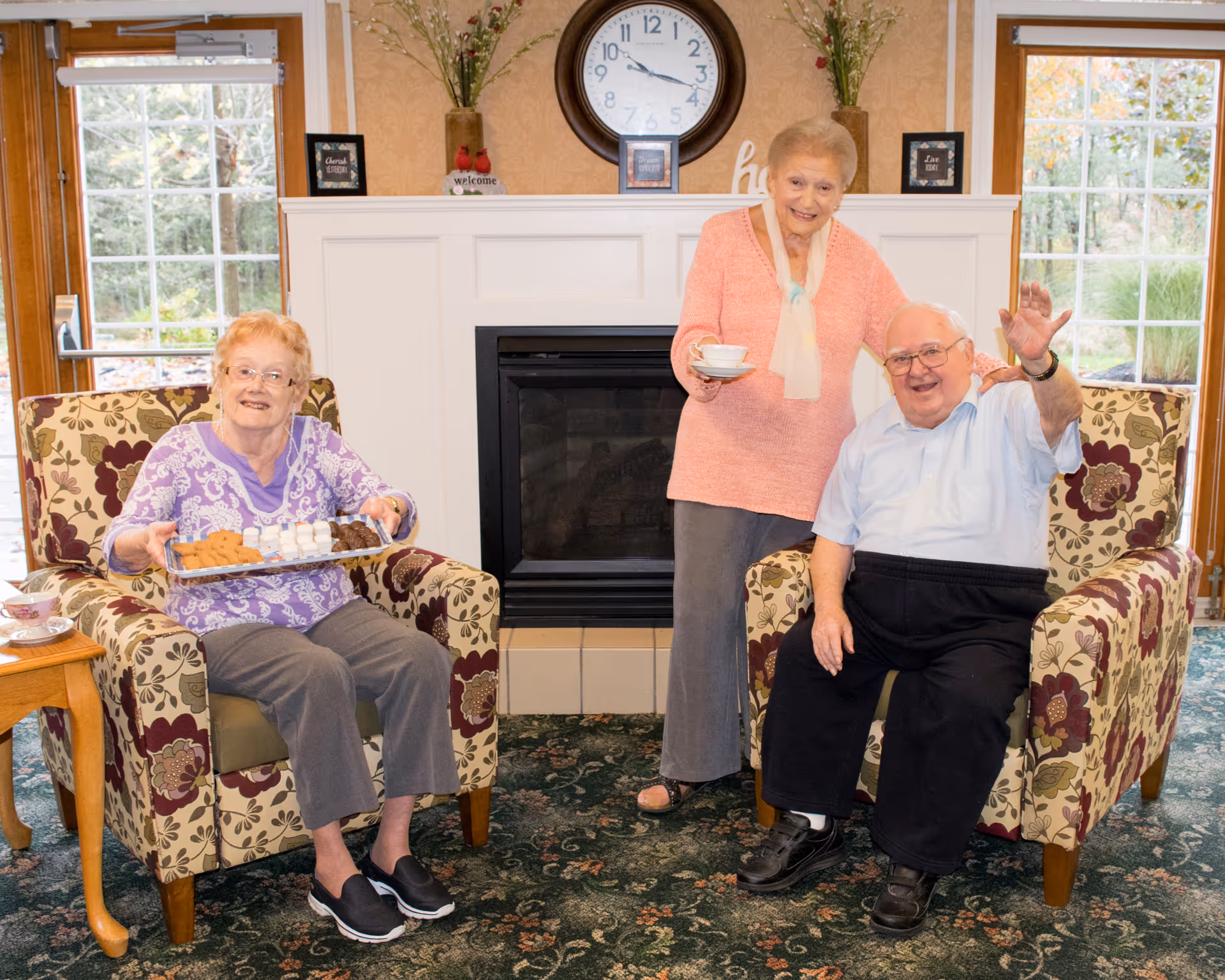 Three elderly people in a cozy living room with floral armchairs and a fireplace. One woman is seated holding a tray of cookies, another woman is standing holding a cup and saucer, and a man is seated waving. Large windows show greenery outside.