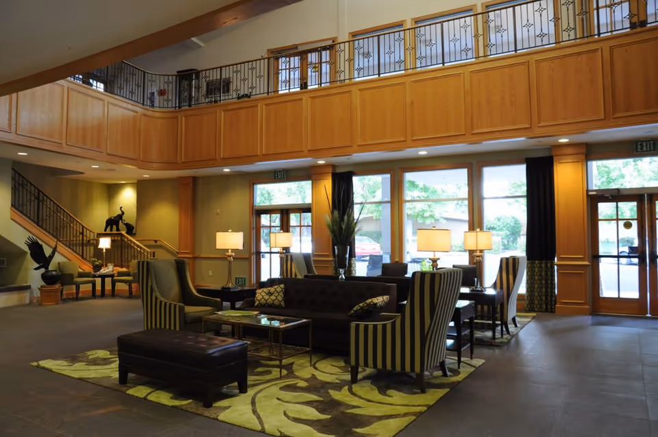 Spacious senior living lobby with sofas, striped armchairs, coffee tables, lamps, large windows and an upstairs balcony.