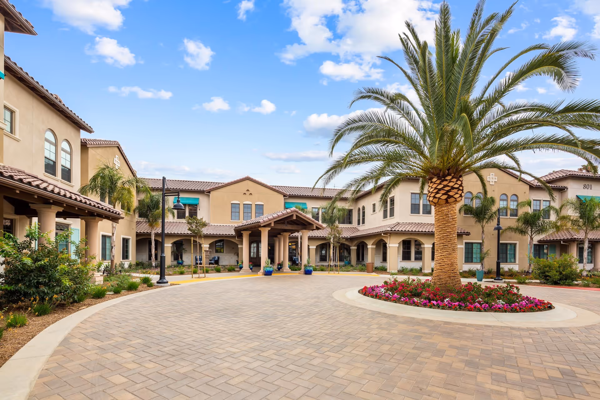 Exterior view of The Kensington Redondo Beach facility featuring a circular driveway with a large palm tree and colorful flowers in the center, surrounded by a beige two-story building with arched windows and a covered entrance under a blue sky with scattered clouds.