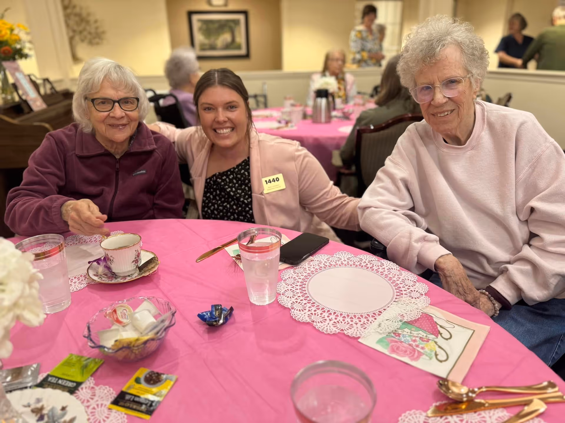 Three women sitting at a table covered with a pink tablecloth in a communal dining area. Two elderly women are seated on either side of a younger woman who is smiling and has her arms around them. The table has cups, glasses of water, tea packets, and utensils. Other people are visible in the background, also seated at tables.