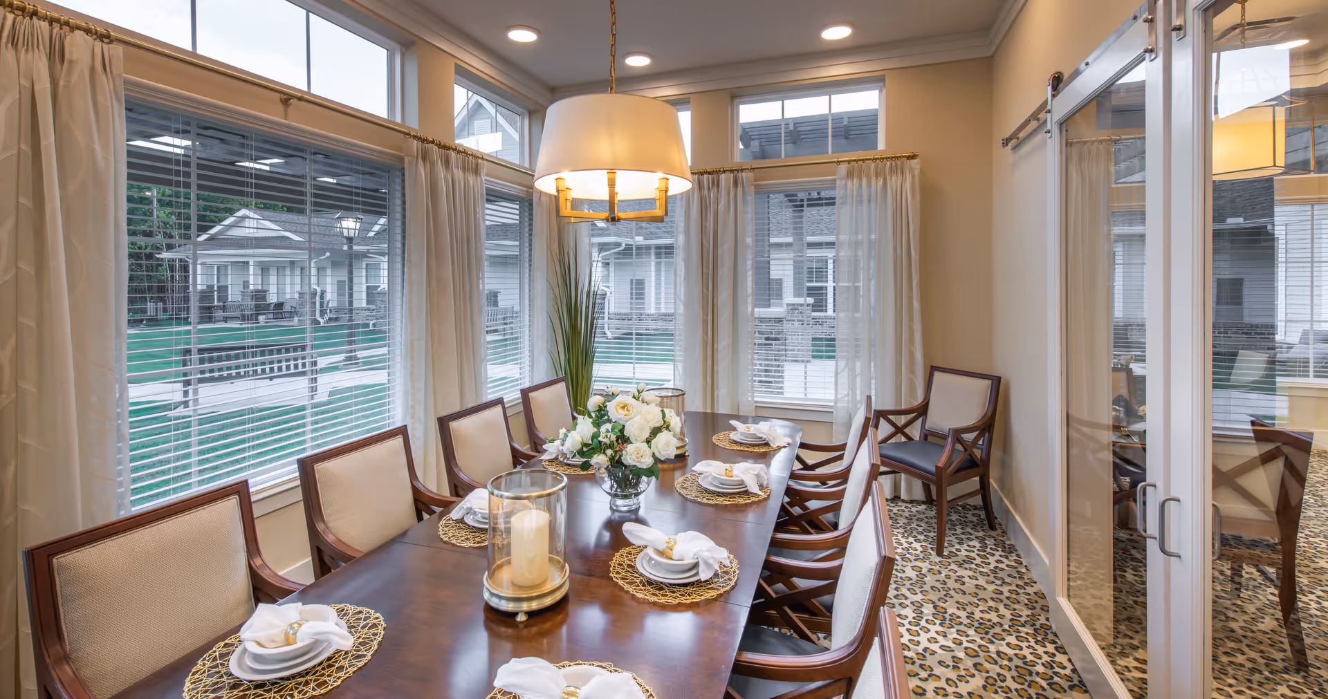 Bright dining room with a long wooden table set for a meal, upholstered chairs, a floral centerpiece, large windows, and a hanging light fixture.