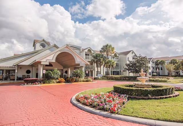 Exterior view of The Palms of Longwood Assisted Living facility showing a large entrance with a covered driveway, landscaped gardens with colorful flowers, palm trees, and a decorative fountain under a partly cloudy sky.