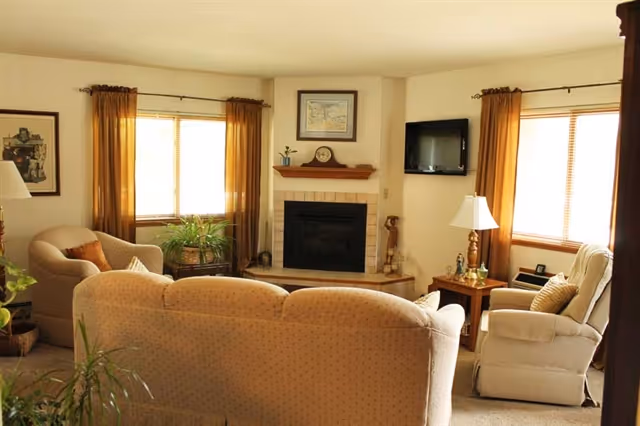A cozy living room with beige upholstered sofas and armchairs arranged around a corner fireplace. The room has two windows with brown curtains, a small wooden side table with a lamp, a wall-mounted TV, and some decorative plants and framed artwork.