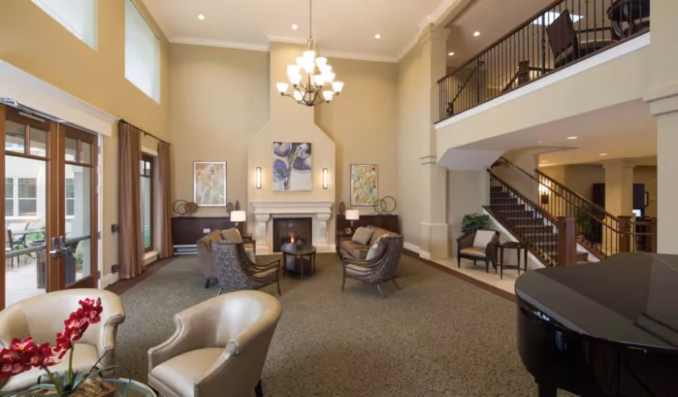 Spacious two-story community lounge with seating arranged around a fireplace, grand piano, staircase and balcony overlooking large windows and glass doors.