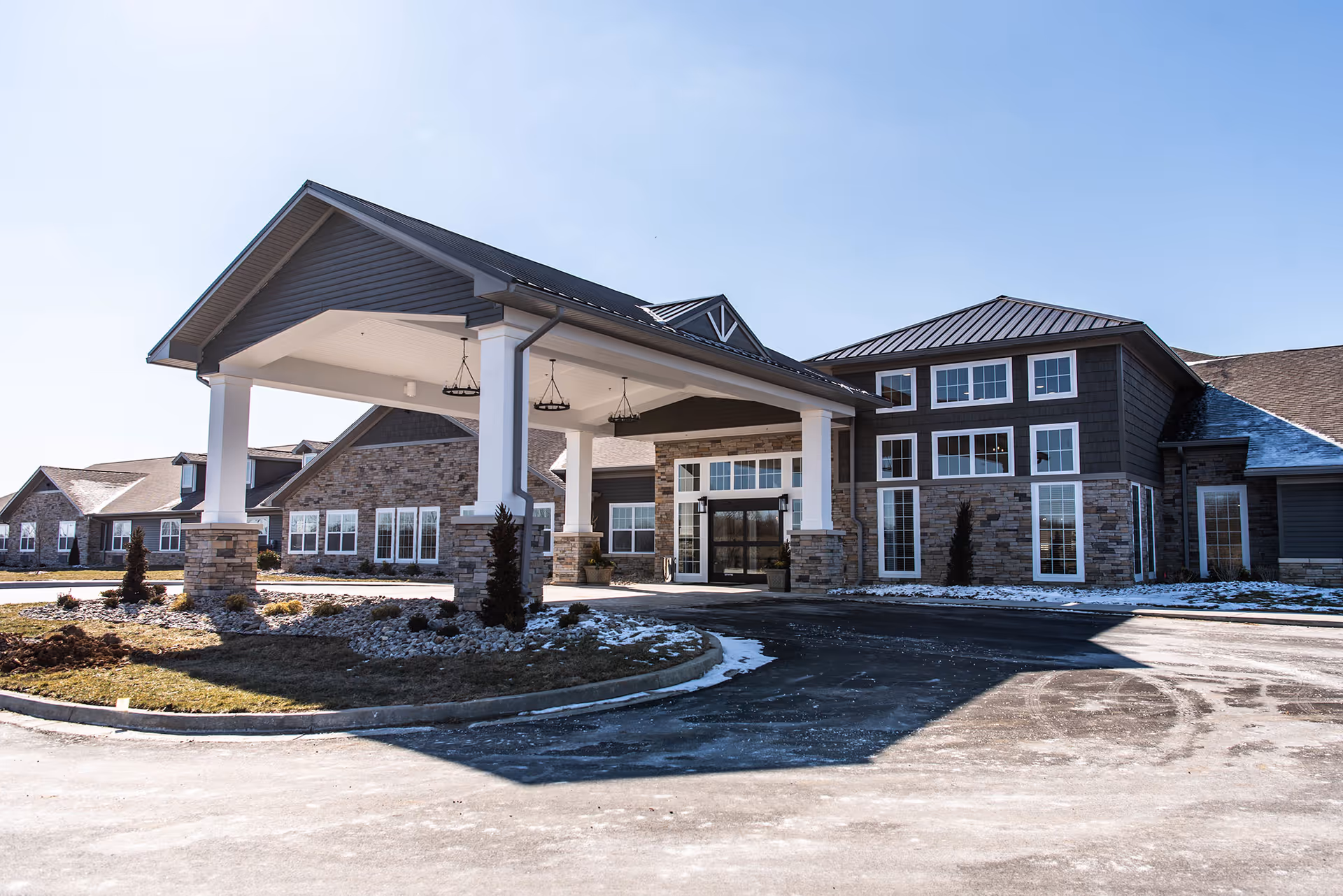 Front exterior of a senior living facility with a covered porte-cochere, stone and siding facade, and light snow on the ground.