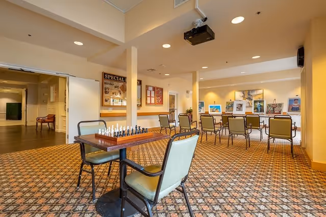 A spacious room with patterned carpet flooring and beige walls, featuring a table with a chessboard and two chairs in the foreground. In the background, there are several chairs arranged around tables displaying framed artwork. The room is well-lit with recessed ceiling lights and has a projector mounted on the ceiling.