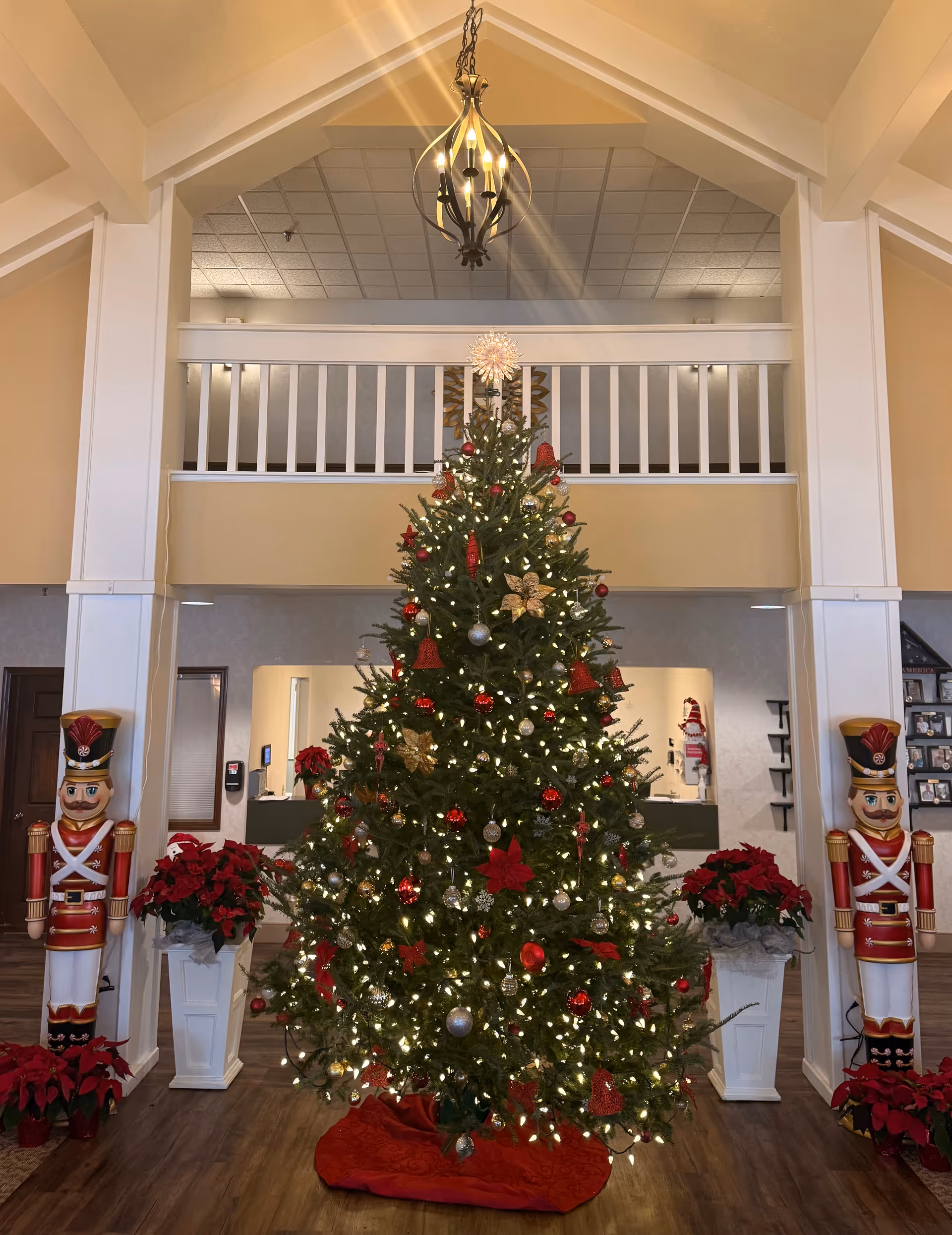 A large decorated Christmas tree with white lights, red and gold ornaments, and a star topper stands in the center of a spacious room with high ceilings. On either side of the tree are tall nutcracker statues and red poinsettia plants in white planters. The room features a wooden floor, white pillars, and a balcony railing above the tree. A chandelier hangs from the ceiling above.