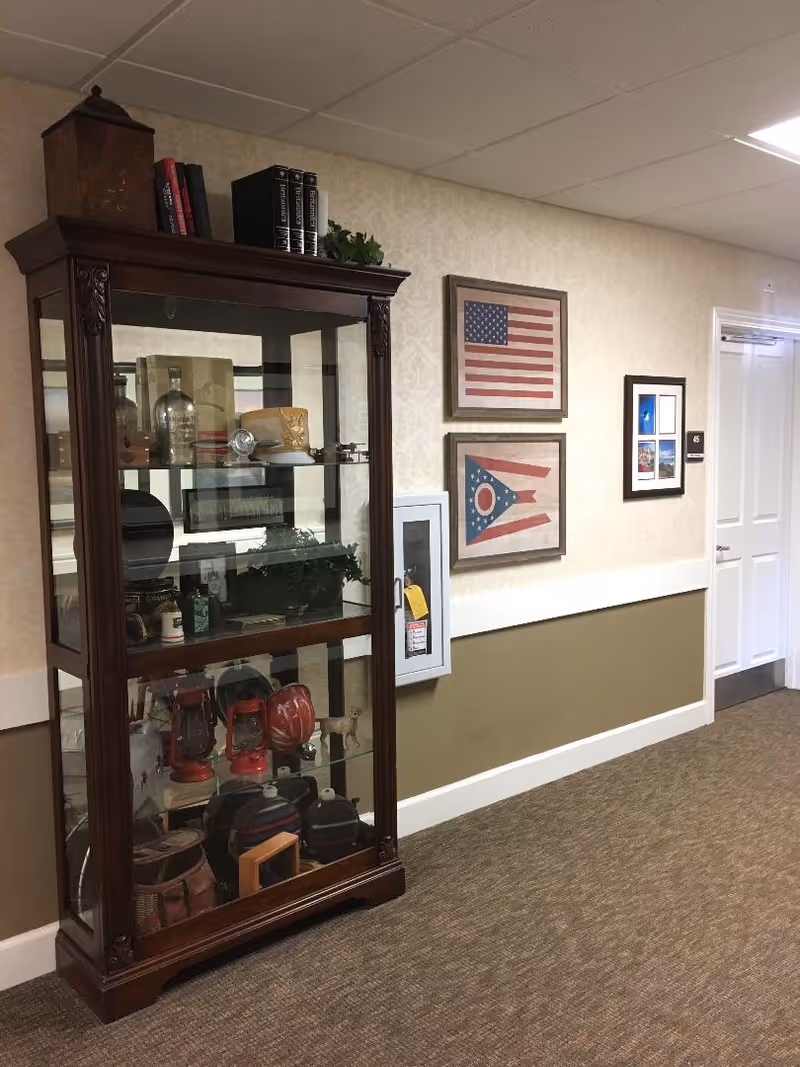 A hallway in an assisted living facility with a wooden display cabinet filled with various decorative items including lanterns, bottles, and books. On the wall next to the cabinet are framed pictures of the American flag and the Ohio state flag, along with another framed photo. The hallway has beige wallpaper with a patterned design on the upper half and a solid olive green color on the lower half, with a white chair rail separating the two. A white door with the number 45 is visible at the end of the hallway.