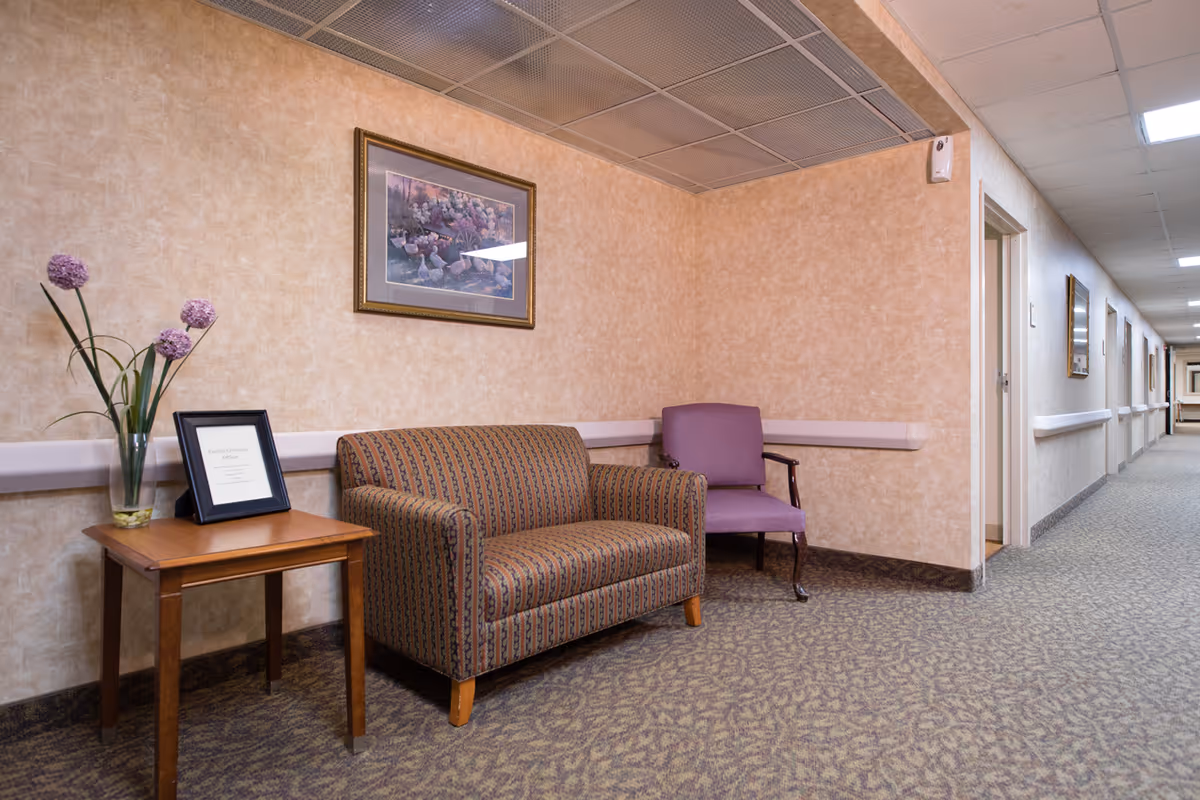 A quiet assisted-living hallway seating area with a patterned loveseat, purple chair, side table with a framed notice and vase of flowers, and a long corridor.