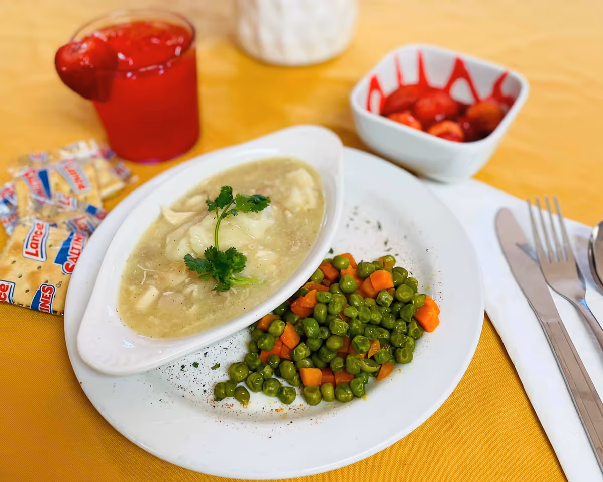 A meal served on a white plate with mashed potatoes topped with gravy and a sprig of parsley, alongside cooked peas and diced carrots. There is a small bowl of strawberries with syrup, a red gelatin dessert, and a pack of Lance saltine crackers on a yellow tablecloth. Silverware is placed on a white napkin to the right of the plate.