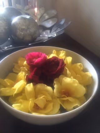 A white bowl of yellow and red rose blossoms sits on a dark tabletop with silver decorative spheres in the background.
