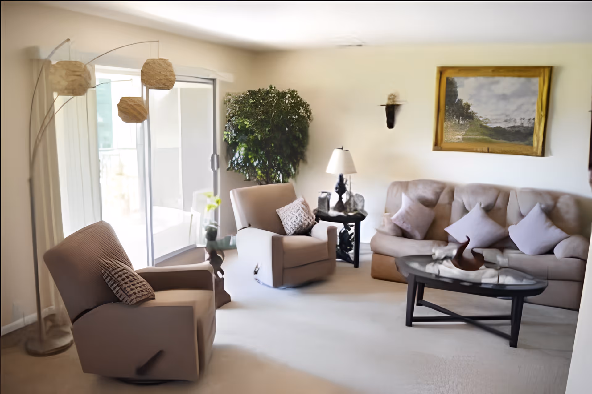 A cozy living room with a beige sofa adorned with four light-colored pillows, two beige armchairs with patterned cushions, a glass coffee table with decorative items, a side table with a lamp, a large potted plant near sliding glass doors, and a floor lamp with three hanging lampshades. A framed landscape painting hangs on the wall.