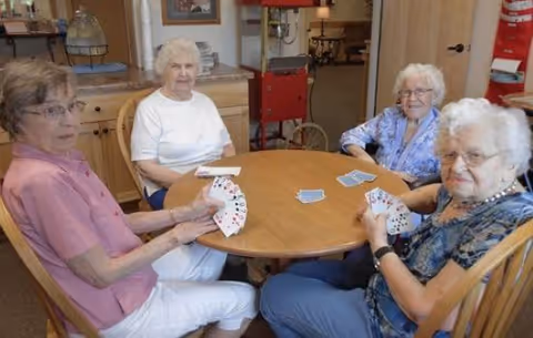Four elderly women sitting around a wooden round table playing cards in a cozy indoor setting with wooden furniture and a fire extinguisher visible in the background.