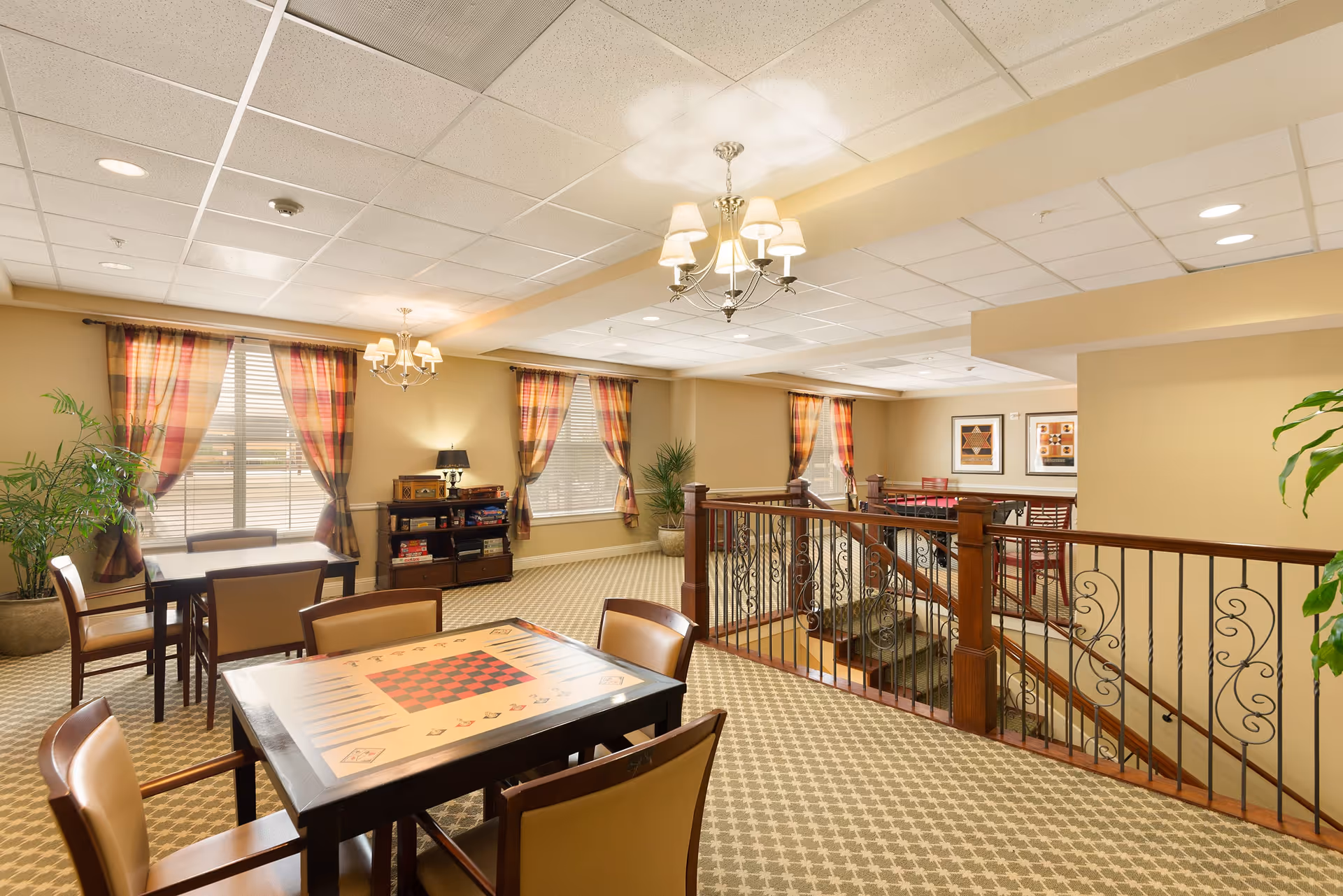 A well-lit common area in a senior living facility featuring multiple tables with chairs, including a game table with a checkerboard design. The room has beige walls, patterned carpet, large windows with plaid curtains, decorative plants, framed artwork on the walls, and a staircase with a wooden and wrought iron railing.