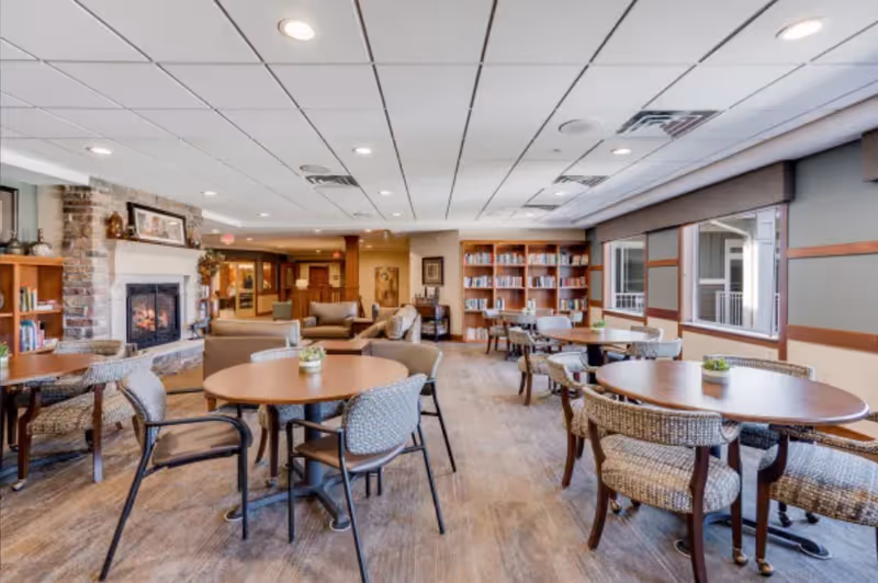 A spacious senior living common area with round tables and chairs arranged for socializing or activities. The room features a cozy stone fireplace on the left, bookshelves filled with books, large windows letting in natural light, and comfortable seating areas with sofas in the background.