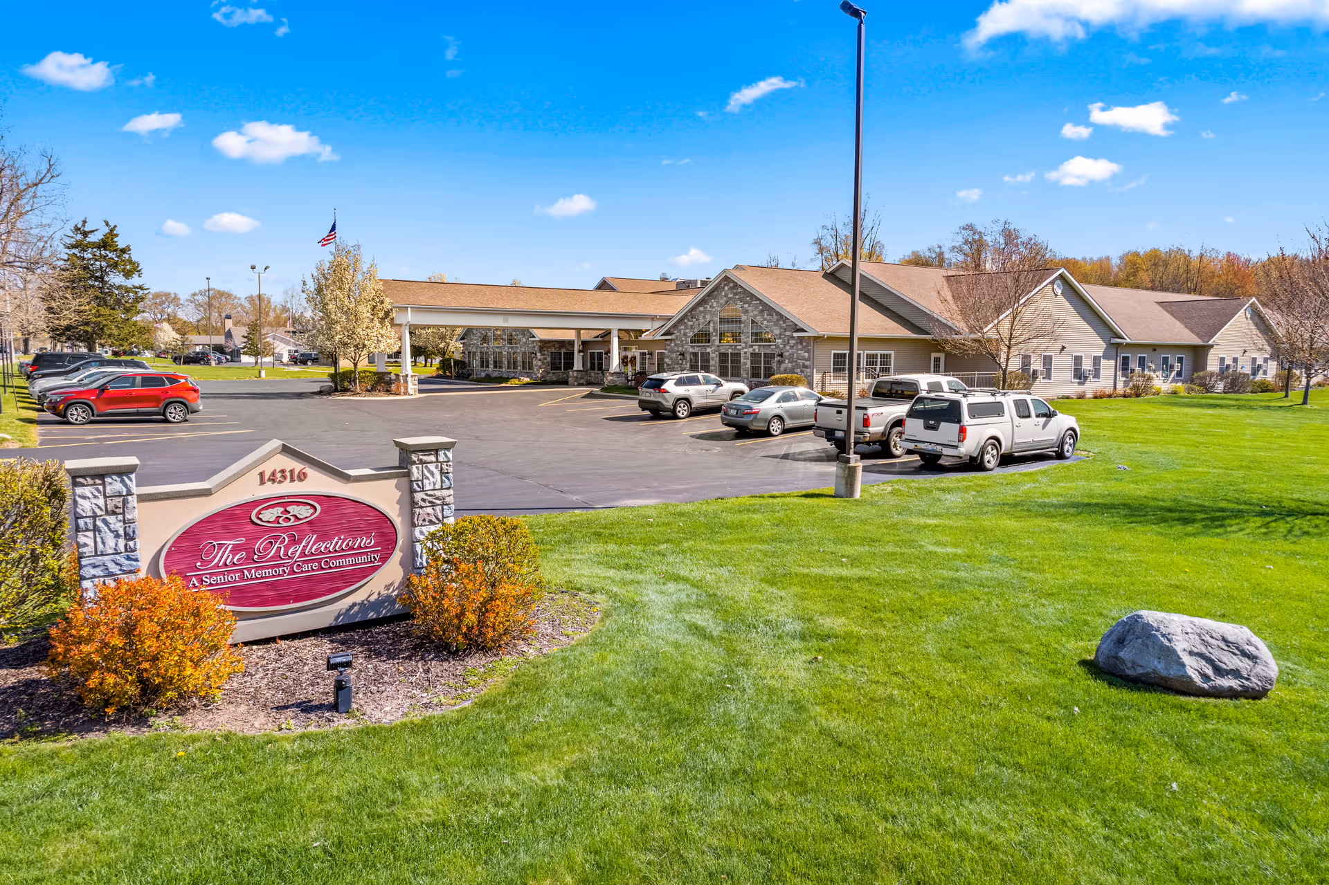 Exterior view of The Reflections memory care building with its entrance sign, parking lot, and well-kept lawn under a blue sky.