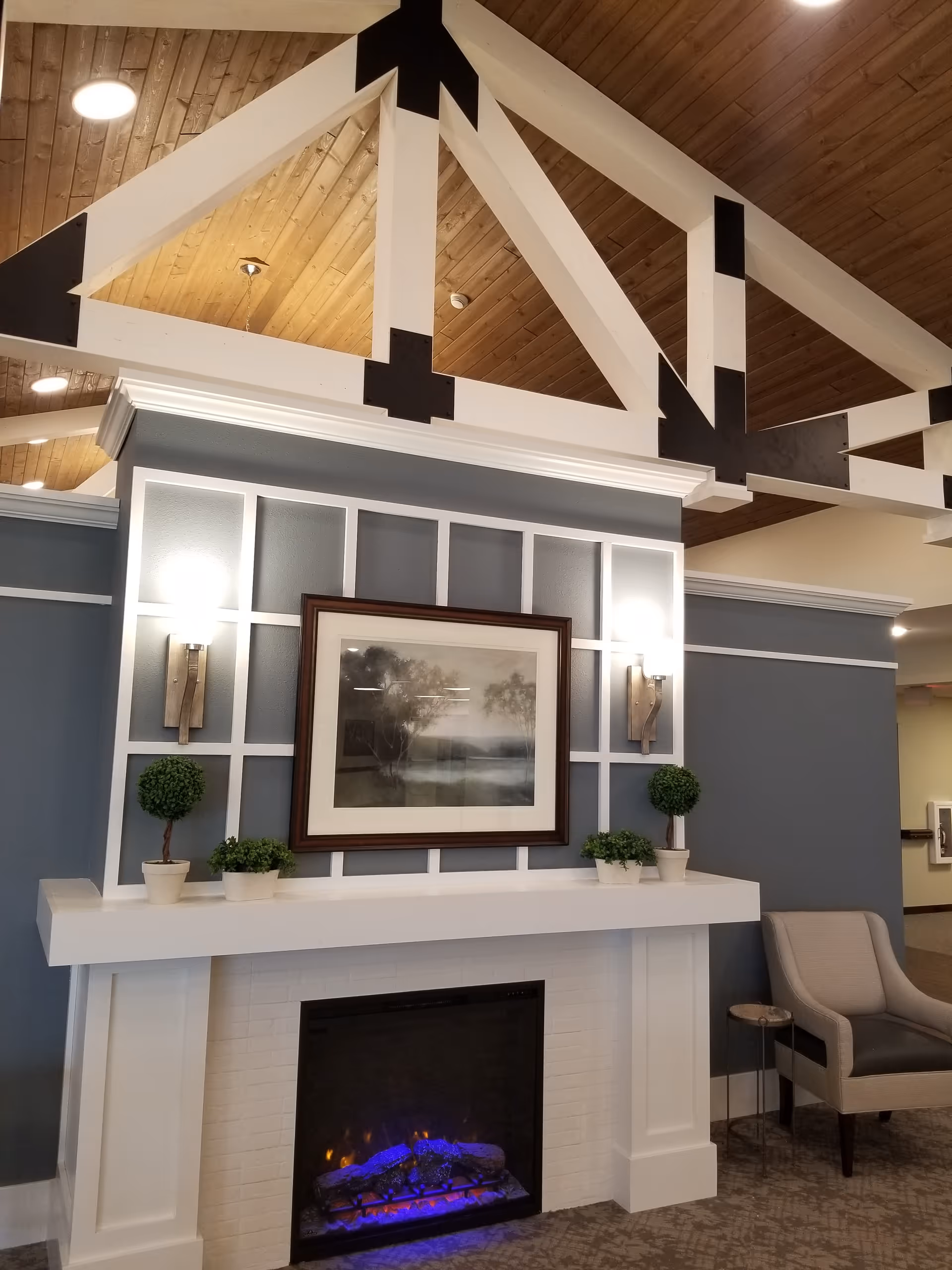 Interior lounge with a white mantel fireplace, framed artwork, potted plants, a chair, and a vaulted wood ceiling with exposed white beams.