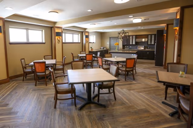 A spacious dining room with multiple square tables and chairs arranged neatly. The room features wood-patterned flooring, beige walls with wooden trim, and several windows letting in natural light. In the background, there is a kitchen area with dark cabinets, a countertop, and a decorative light fixture hanging from the ceiling.