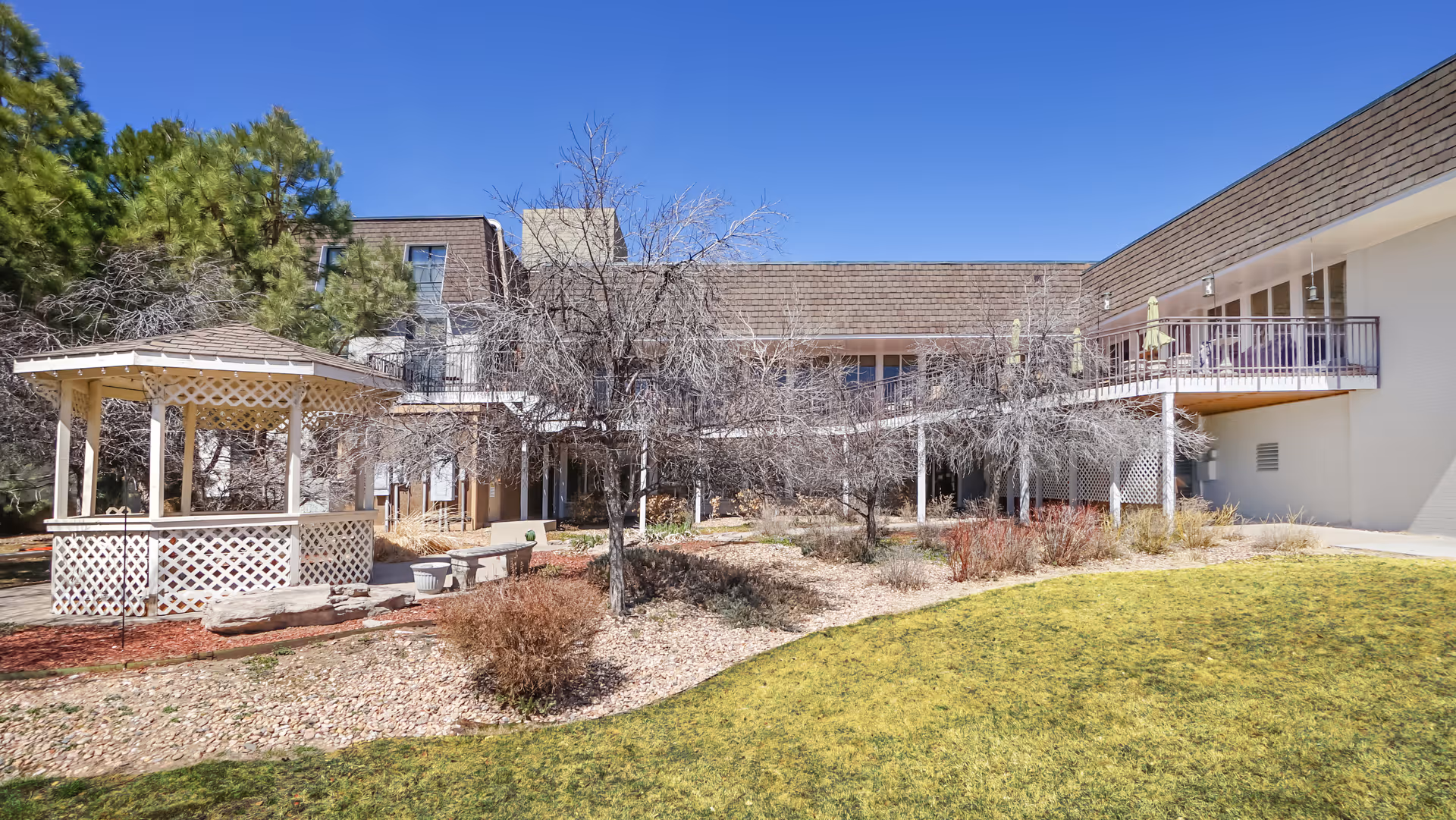 Outdoor view of a senior living facility with a white gazebo, leafless trees, landscaped garden beds with shrubs, and a two-story building with balconies and patio umbrellas under a clear blue sky.