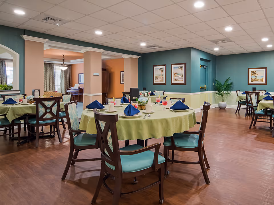 Dining room in The Windsor of Palm Coast facility with round tables covered in green tablecloths, set with blue napkins, cups, and glasses. The room has wooden chairs with green cushions, teal and beige walls, framed artwork, and a wooden floor. There is a piano visible in an adjacent room.