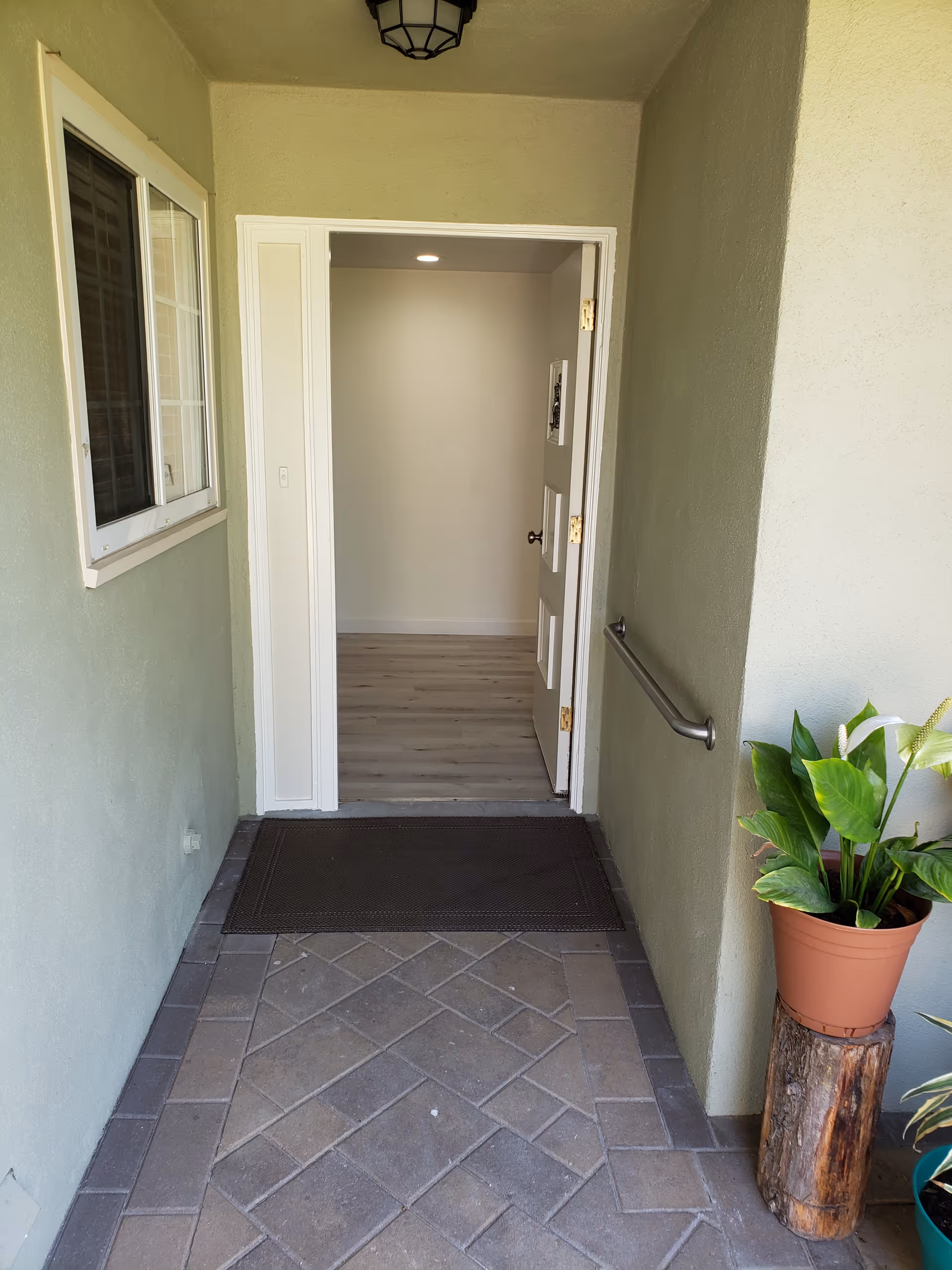 Entrance hallway with a tiled floor leading to an open white door. There is a black doormat in front of the door, a window on the left wall, a handrail on the right wall, and a potted plant on a wooden stand to the right.