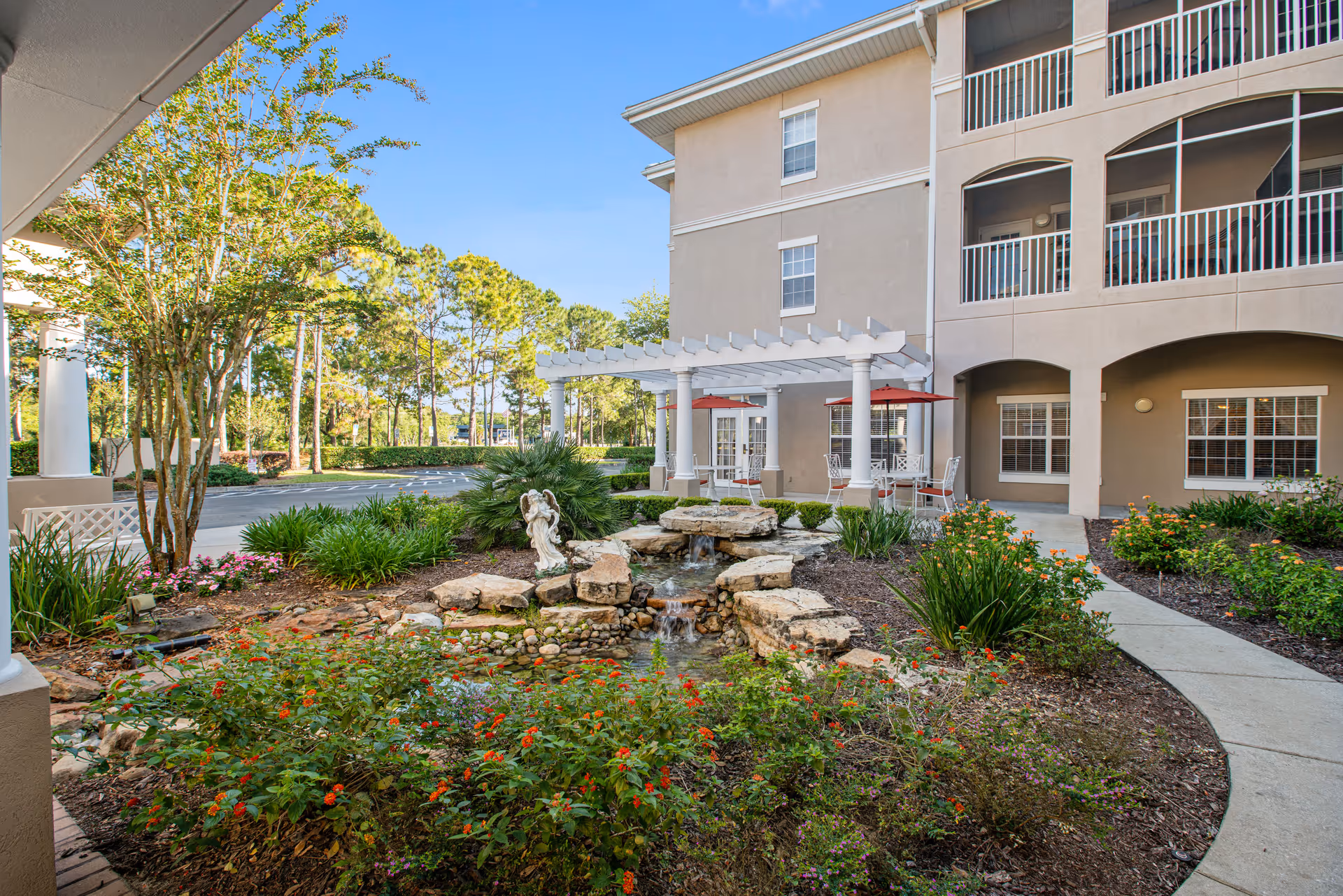 Outdoor garden area at The Residence at Timber Pines featuring a small pond with rocks and a water fountain, surrounded by various plants and flowers. There is a white pergola with outdoor seating and red umbrellas next to a beige multi-story building with balconies and windows.