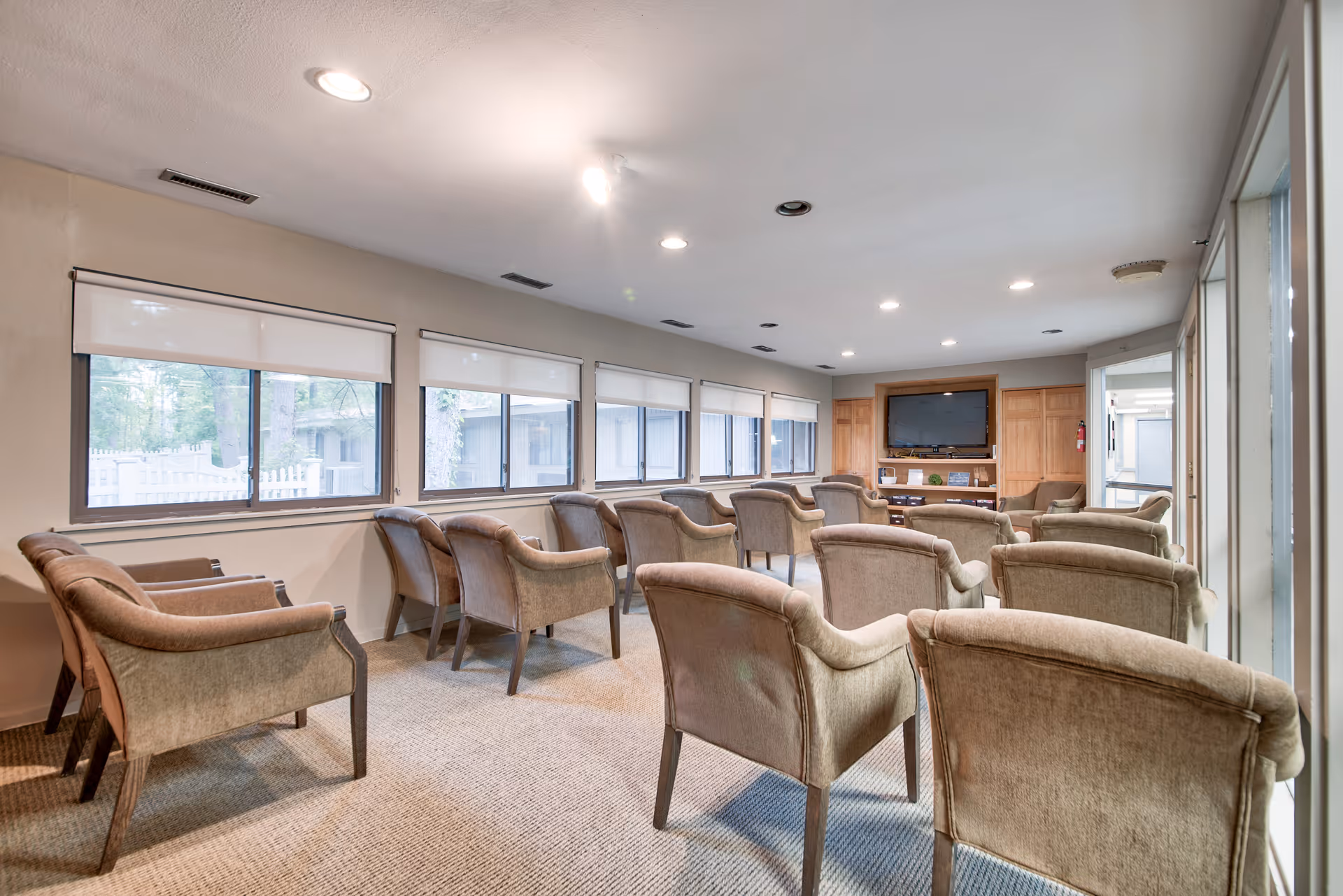 A well-lit common room with multiple beige upholstered armchairs arranged in rows facing a large flat-screen TV mounted on a wooden cabinet. The room has several windows with white roller shades, beige walls, and a carpeted floor.