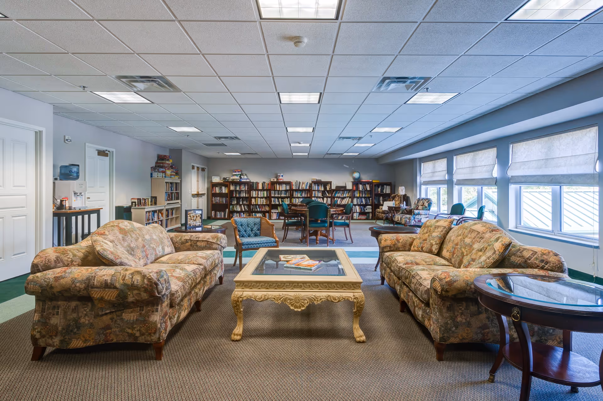 A spacious common area in a senior living facility featuring two patterned sofas facing each other with a glass-top coffee table in between. Behind the seating area are bookshelves filled with books and several tables with chairs. Large windows with white blinds allow natural light to fill the room.
