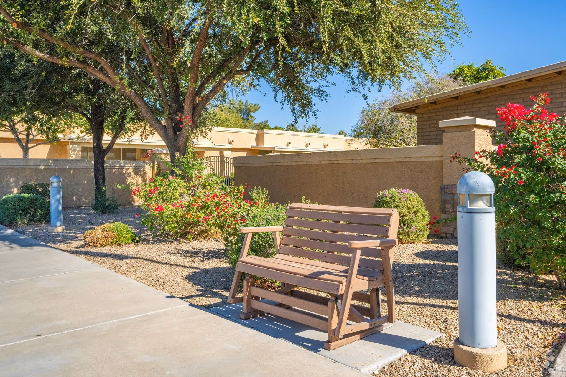 Outdoor seating area with a wooden bench on a concrete path surrounded by desert landscaping including bushes, flowering plants, and trees under a clear blue sky.