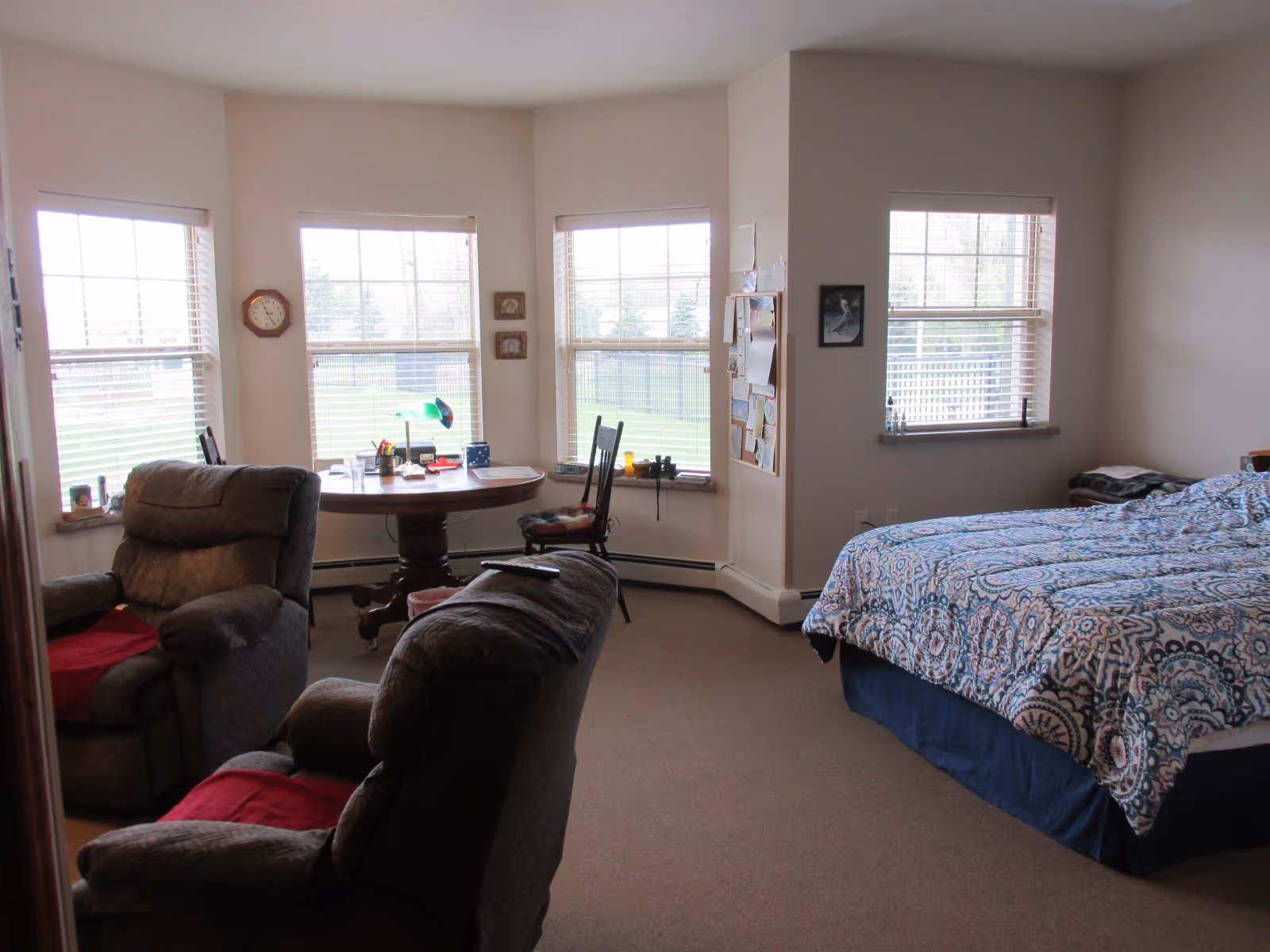 Bright studio bedroom featuring a patterned bed, two recliners, and a round table by bay windows.