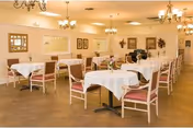 A spacious dining room with multiple tables covered in white tablecloths, each surrounded by wooden chairs with pink cushions. The room is well-lit with chandeliers hanging from the ceiling and decorated with framed pictures and floral arrangements on the walls.