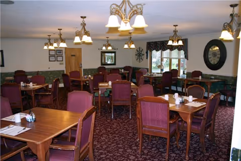 A dining room with multiple wooden tables and chairs upholstered in maroon fabric. The tables are set with white napkins, cups, and condiments. The room has patterned carpet, several hanging light fixtures, a large clock on the wall, and windows with valances letting in natural light.