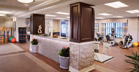 Interior view of a rehabilitation and post-acute health care facility showing a reception desk with decorative plants and sculptures. In the background, there is exercise equipment including stationary bikes and weights, with large windows covered by striped curtains allowing natural light into the room.