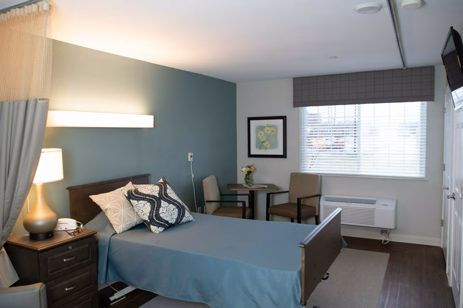 A neatly arranged bedroom in a senior living facility with a single bed covered in a blue bedspread and two decorative pillows. There is a bedside table with a lamp and a telephone, a small round table with two chairs, a framed floral picture on the wall, a window with blinds, and an air conditioning unit below the window. The walls are painted in soft blue and white tones.