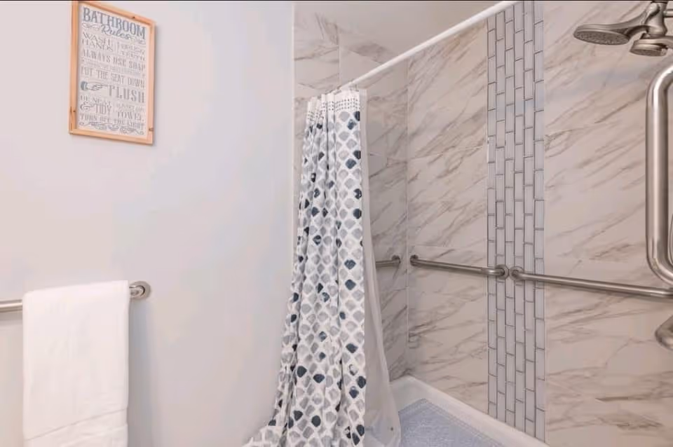 Bathroom with a white towel hanging on a metal bar, a shower area with marble-patterned tiles and a vertical strip of smaller rectangular tiles, a shower curtain with a blue and gray geometric pattern, and a framed sign on the wall with bathroom rules.
