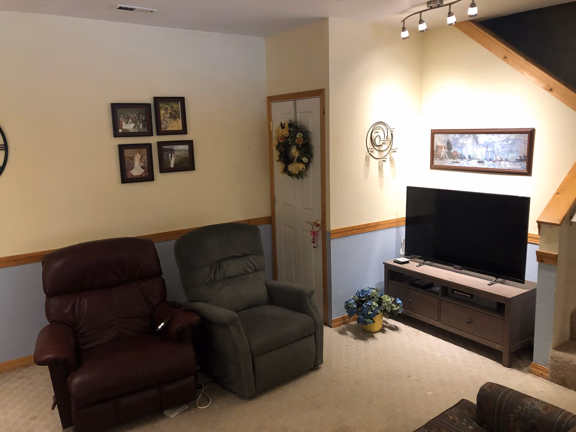 A cozy living room with two recliner chairs, one brown leather and one green fabric, positioned near a small white door decorated with a floral wreath. A flat-screen TV sits on a wooden TV stand with drawers, and a potted plant with blue flowers is placed on the floor beside it. The walls are painted cream and light blue with wooden trim, and there are framed pictures and a decorative wall clock hanging on the walls. A staircase with wooden railing is visible on the right side.