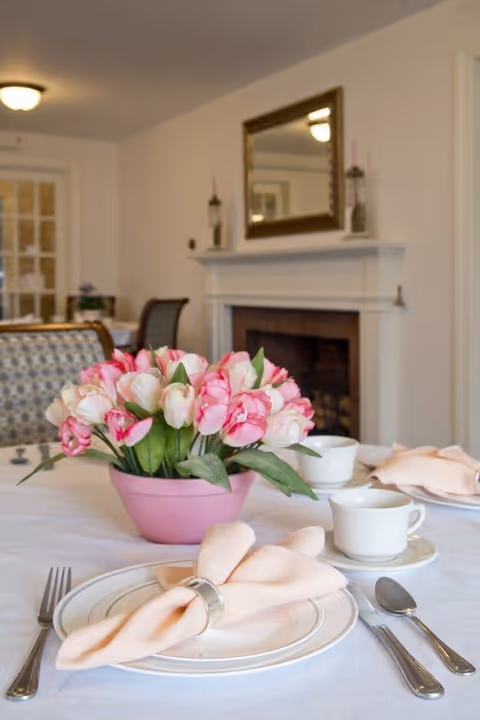 A dining table set with white plates, silver cutlery, and peach-colored napkins held by napkin rings. A pink pot with pink and white tulips is placed in the center of the table. In the background, there is a fireplace with a mirror above it and a chair with patterned upholstery.