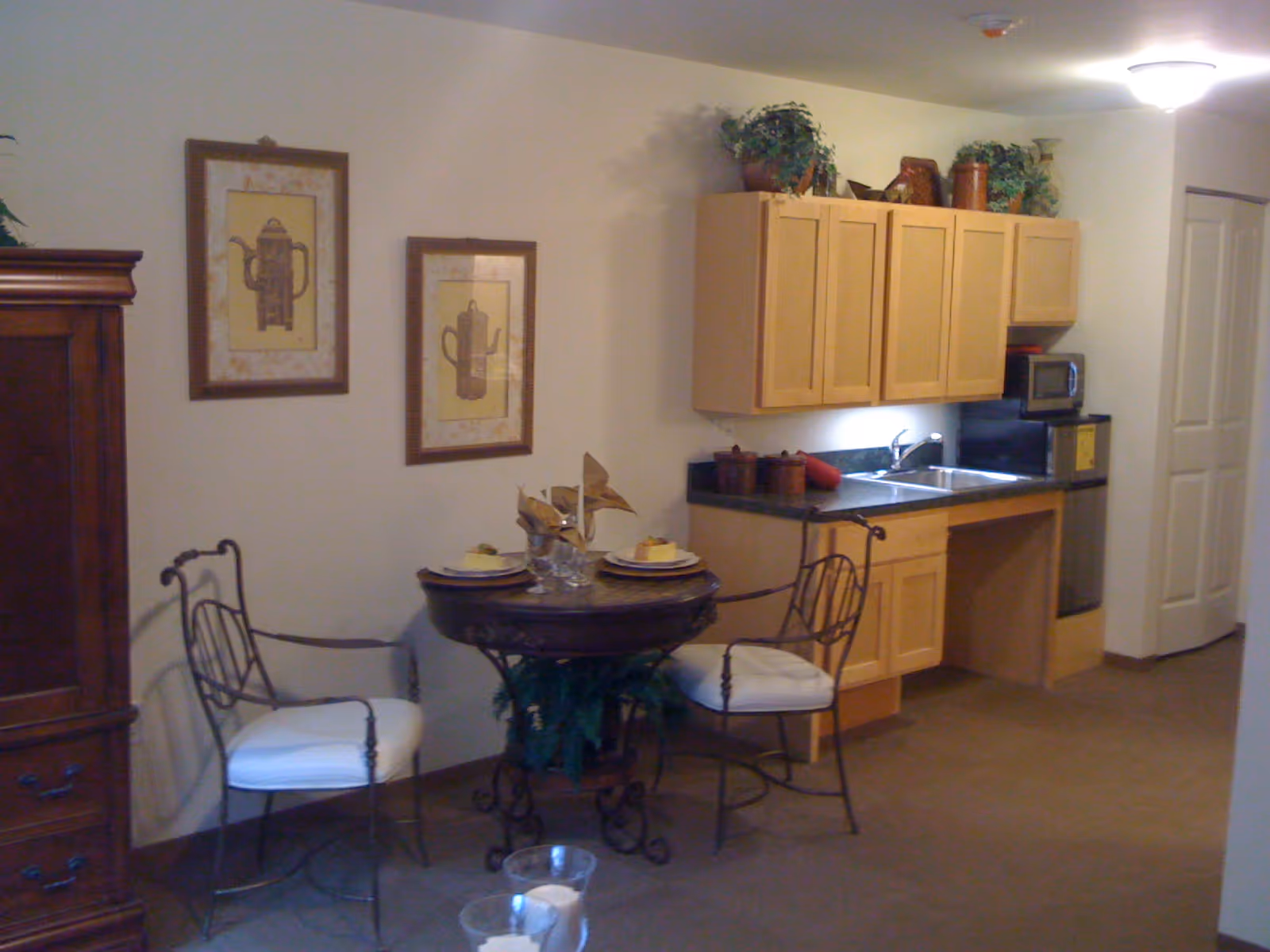 A small kitchenette area with light wood cabinets, a double sink, a microwave, and a mini refrigerator. In front of the kitchenette is a round wooden table set for two with plates, glasses, and napkins, accompanied by two metal chairs with white cushions. Two framed pictures of coffee pots hang on the wall above the table. There are decorative plants on top of the cabinets and under the table. The room has beige walls and carpeted floor.