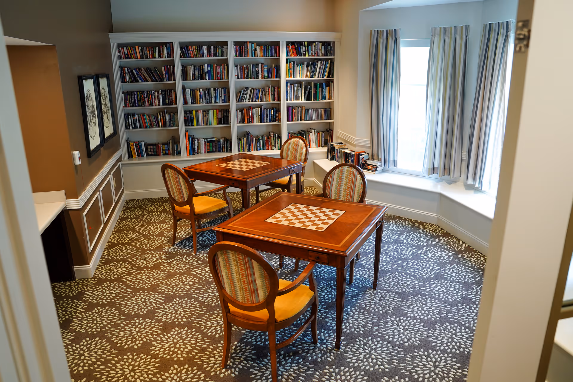 A cozy reading and game room with two wooden tables featuring built-in chessboards, surrounded by four upholstered chairs with striped cushions. A large bookshelf filled with books lines the back wall, and a window with striped curtains lets in natural light. The room has patterned carpet and warm, neutral-colored walls.