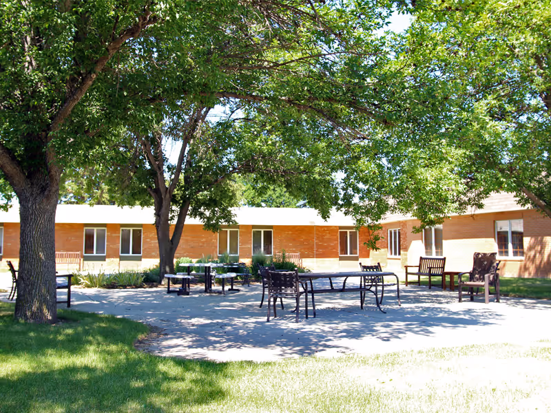 Outdoor patio area with several metal and wooden chairs and tables arranged under large leafy trees, adjacent to a single-story brick building with multiple windows.
