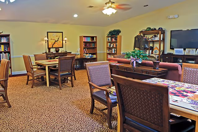 A cozy common area in a senior living facility featuring multiple wooden tables and chairs, bookshelves filled with books, a ceiling fan with lights, a large flat-screen TV mounted on the wall, and decorative plants on a wooden console table. The room has warm yellow walls and patterned carpet flooring.