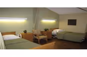 A nursing facility room with two single beds, each with green bedding and white pillows. Between the beds are two wooden nightstands with small potted plants on top. A striped armchair is placed between the nightstands. The walls are light-colored, and there are two wall-mounted lights above each bed providing illumination.