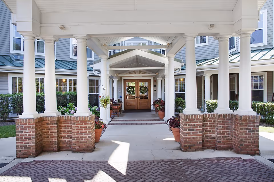 Covered portico with white columns and brick bases leading to double wooden entrance doors flanked by potted plants.