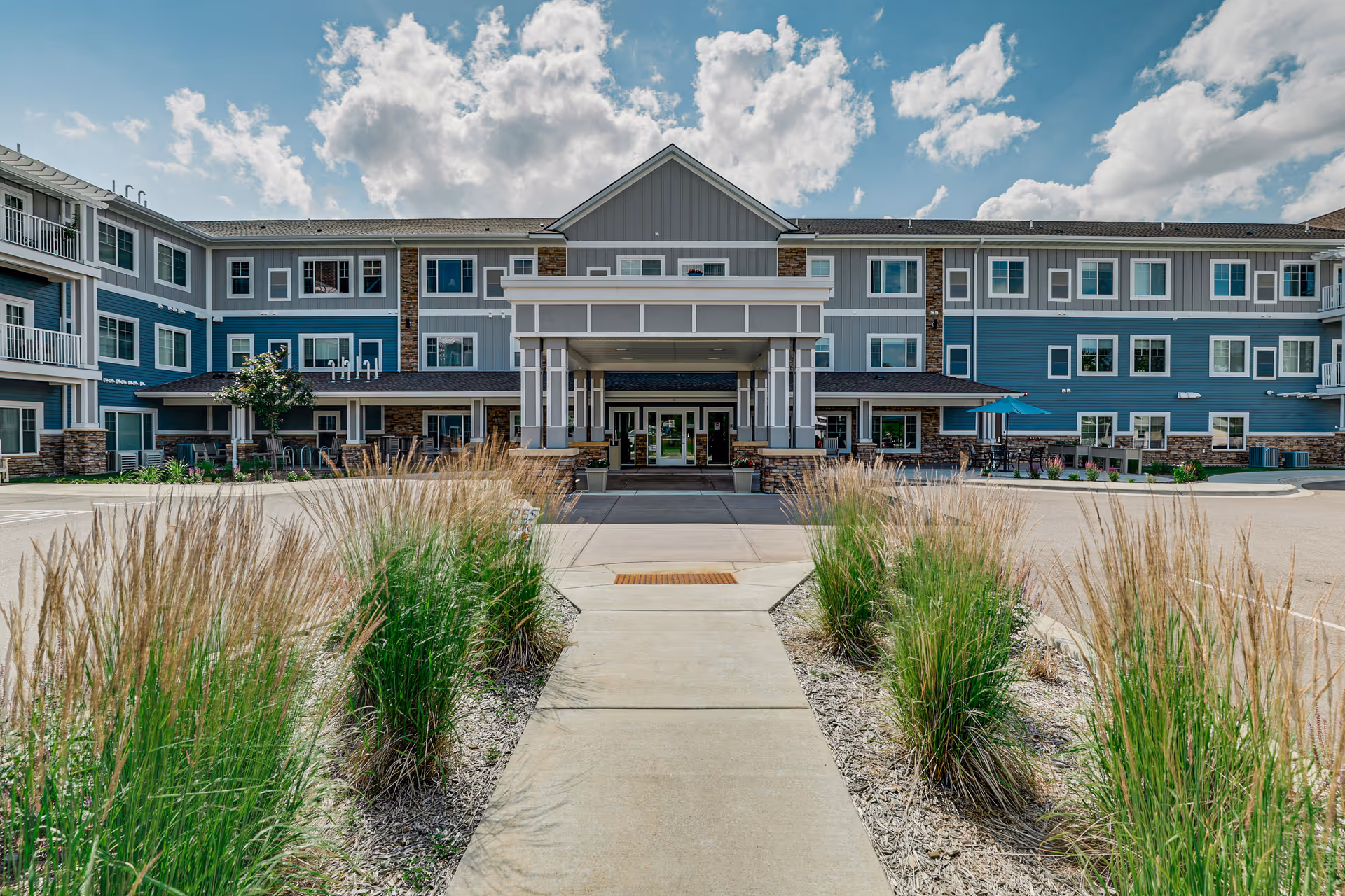 Front entrance of a three-story senior living building with a covered drop-off, walkway, and ornamental grasses.