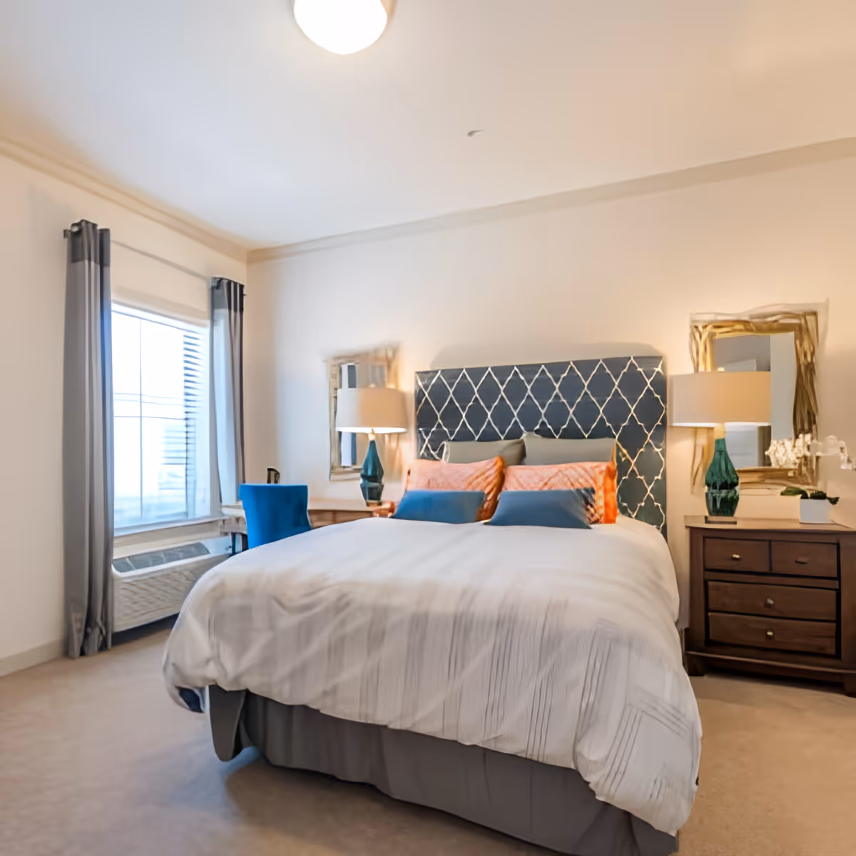 A well-lit bedroom with a large bed featuring a dark blue headboard with a diamond pattern. The bed is made with white bedding and decorated with orange and blue pillows. On either side of the bed are wooden nightstands with matching lamps and decorative mirrors above them. A window with gray curtains and a blue chair are visible on the left side of the room.
