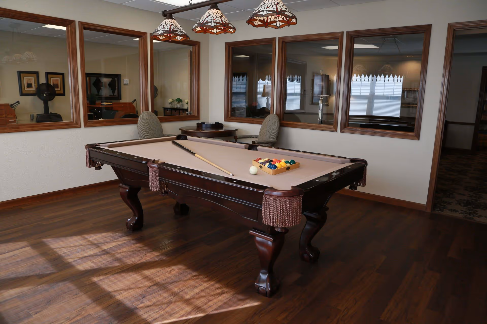 A room with a pool table set up for a game, featuring a cue stick and a rack of billiard balls. The room has wooden flooring, beige walls, and several large windows with wooden frames. There are two upholstered chairs and a small round table in the corner. Three decorative pendant lights hang above the pool table.