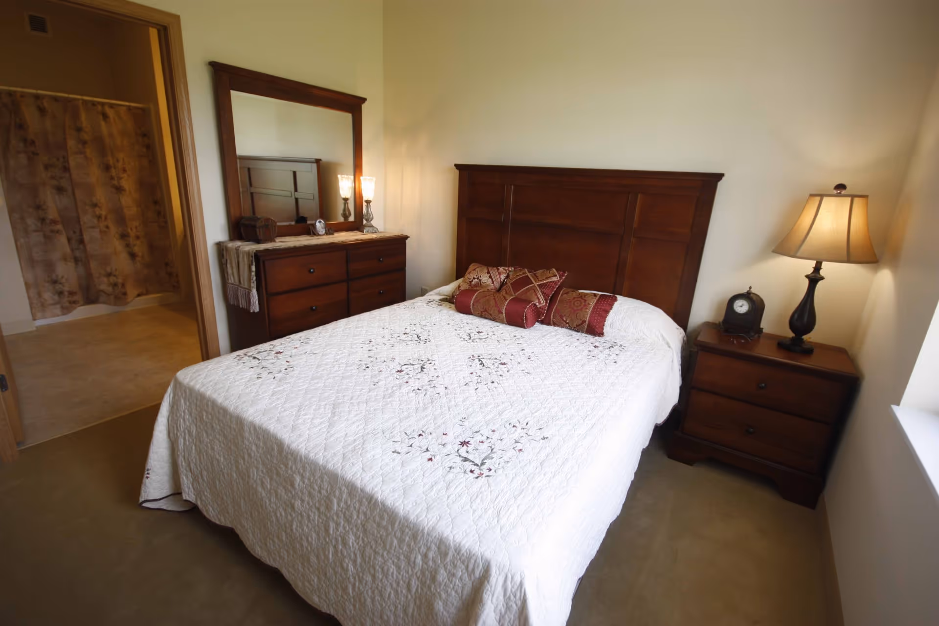 A cozy bedroom with a wooden bed frame and a white quilted bedspread decorated with floral patterns. There are several decorative pillows on the bed. To the right of the bed is a wooden nightstand with a classic lamp and a small clock. On the left side of the room is a wooden dresser with a large mirror and two lit lamps. An open doorway reveals a bathroom with a floral shower curtain.
