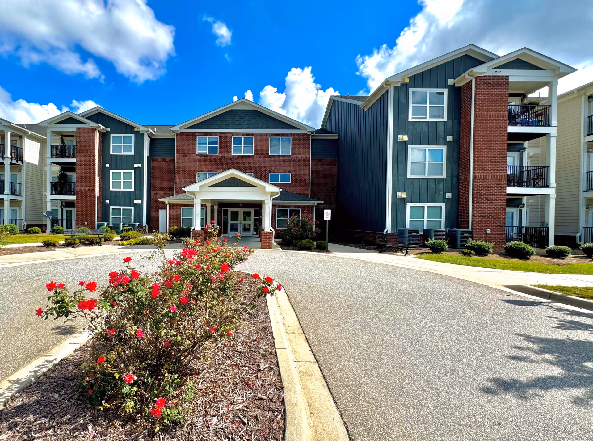 Front entrance of a three-story senior living building with a circular driveway and red flowers in the foreground.